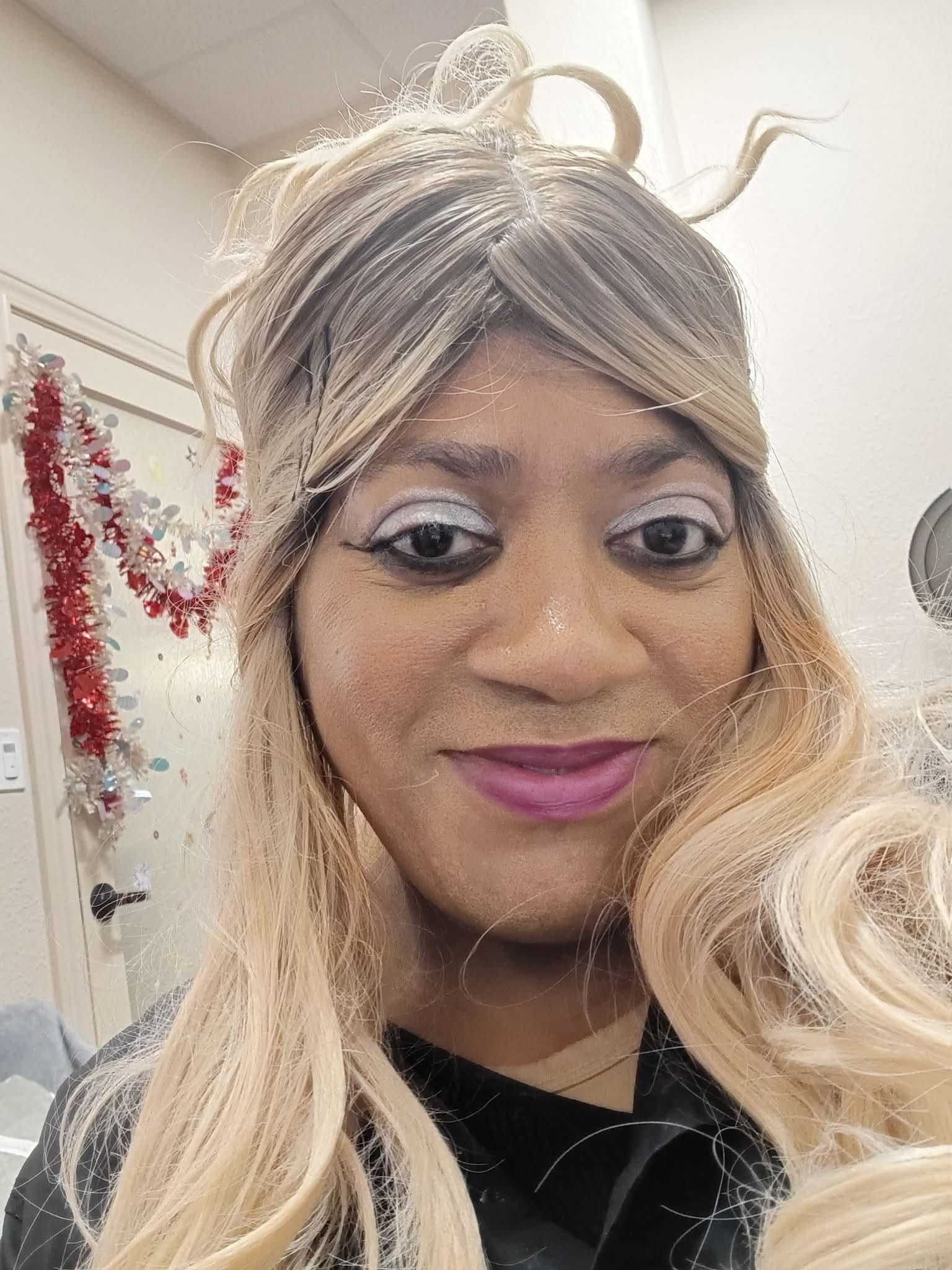 Person with long blonde hair and purple lipstick smiling in a decorated room.