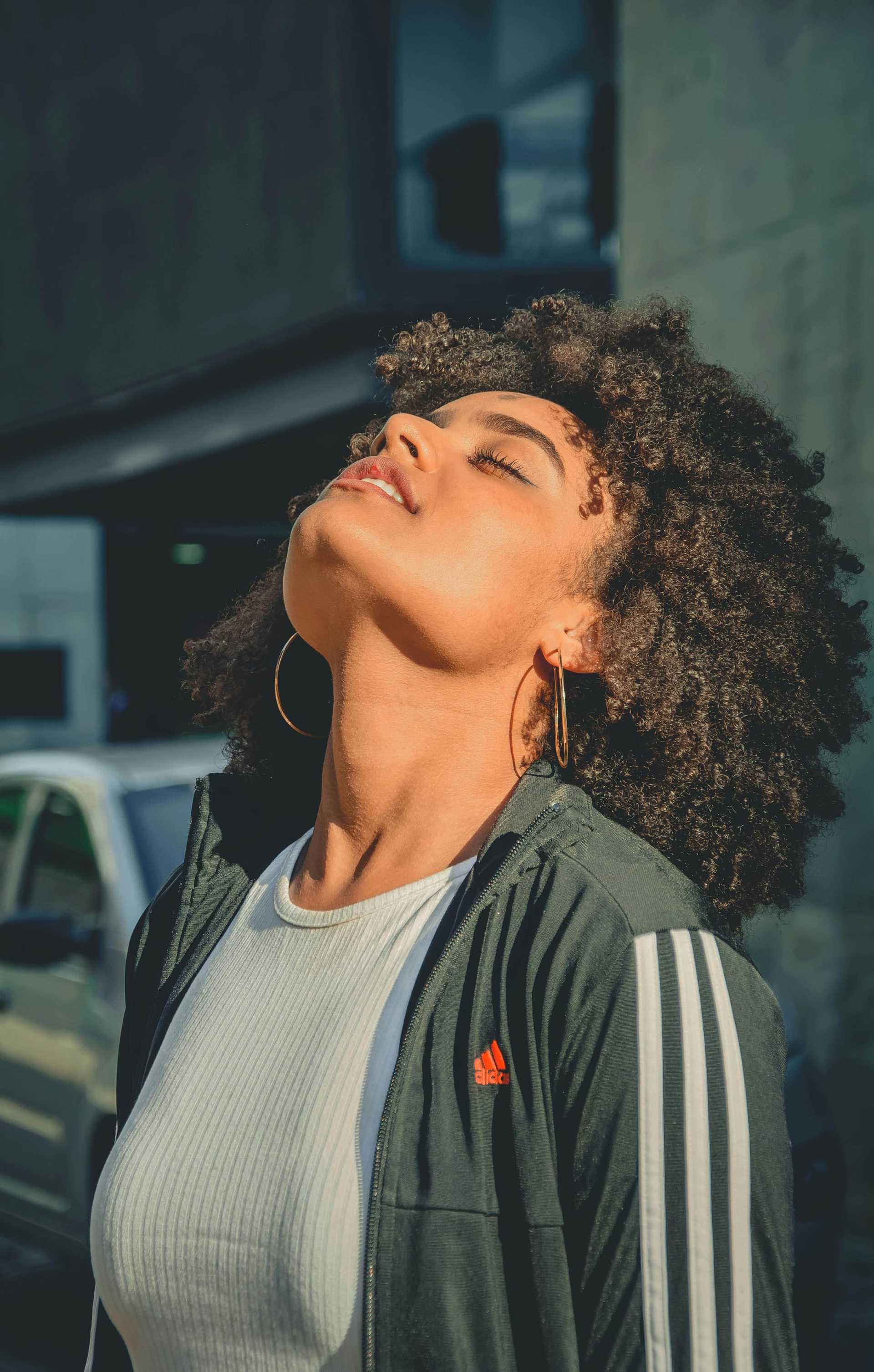 Woman enjoys sunlight, eyes closed, wearing a black jacket with Adidas logo.